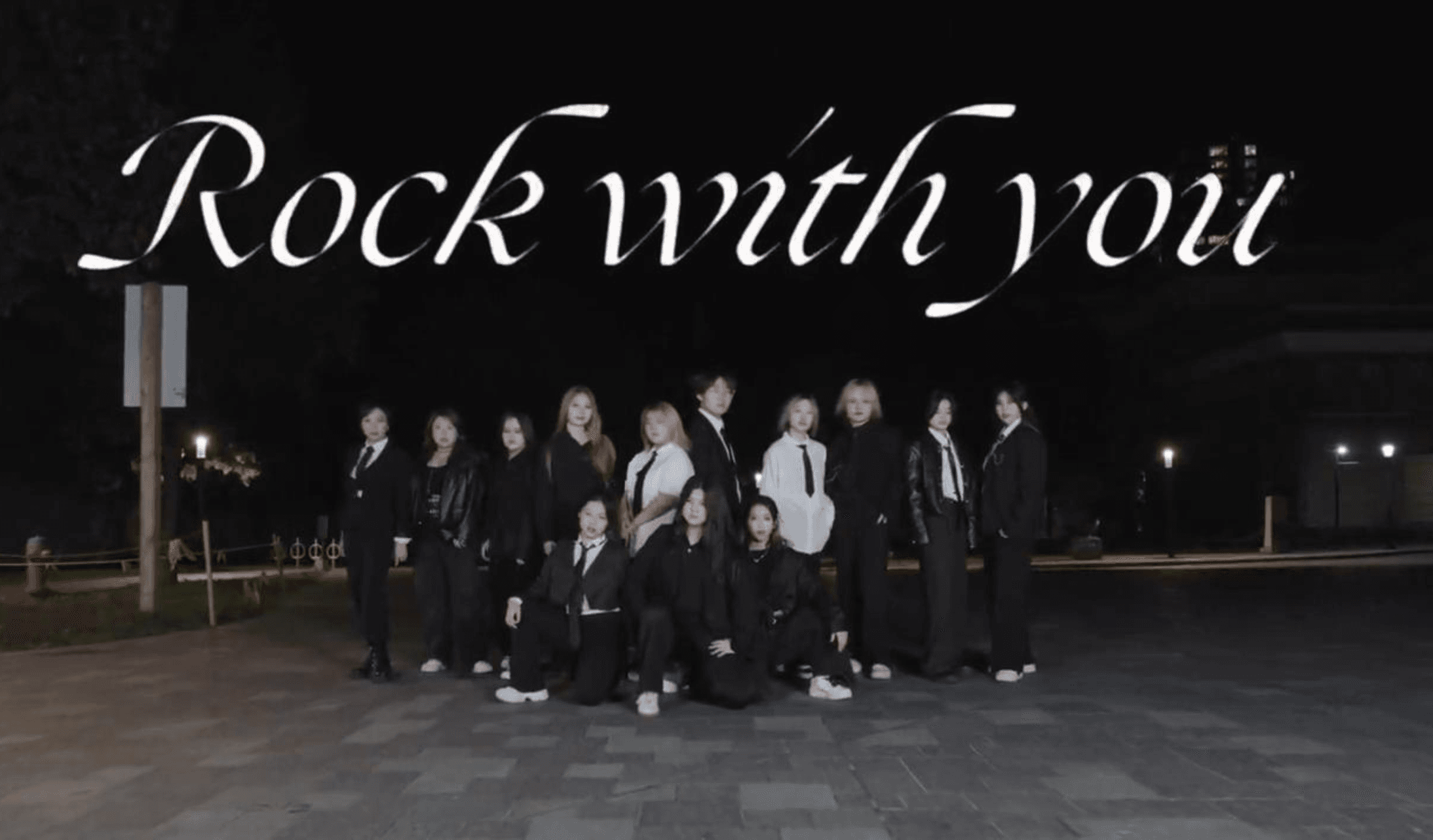 Thirteen dancers in black and white outfits posing outdoors at night for Rock With You by Seventeen.