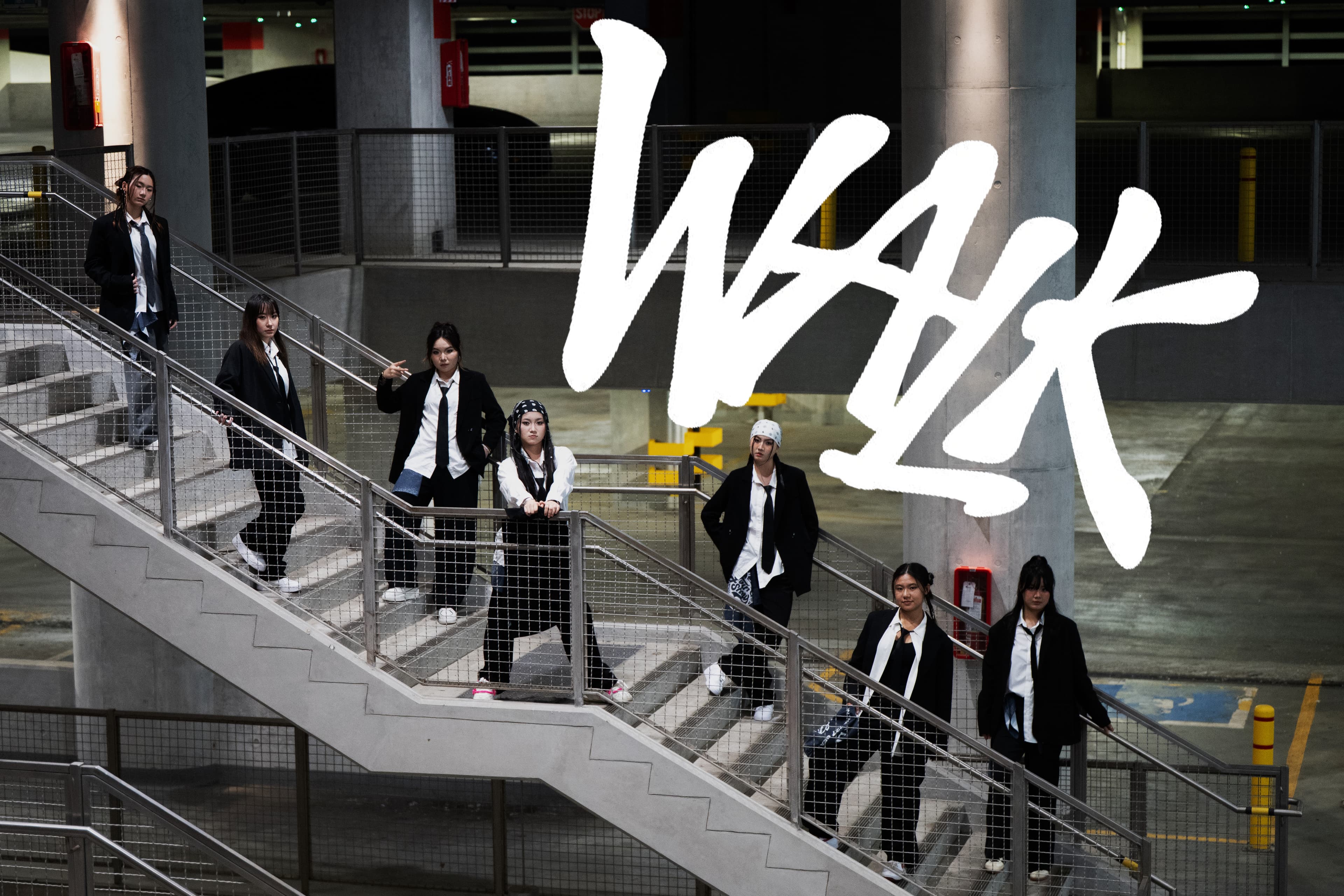 Seven dancers in black suits and white shirts posing on a concrete parking garage staircase for Walk by NCT 127.